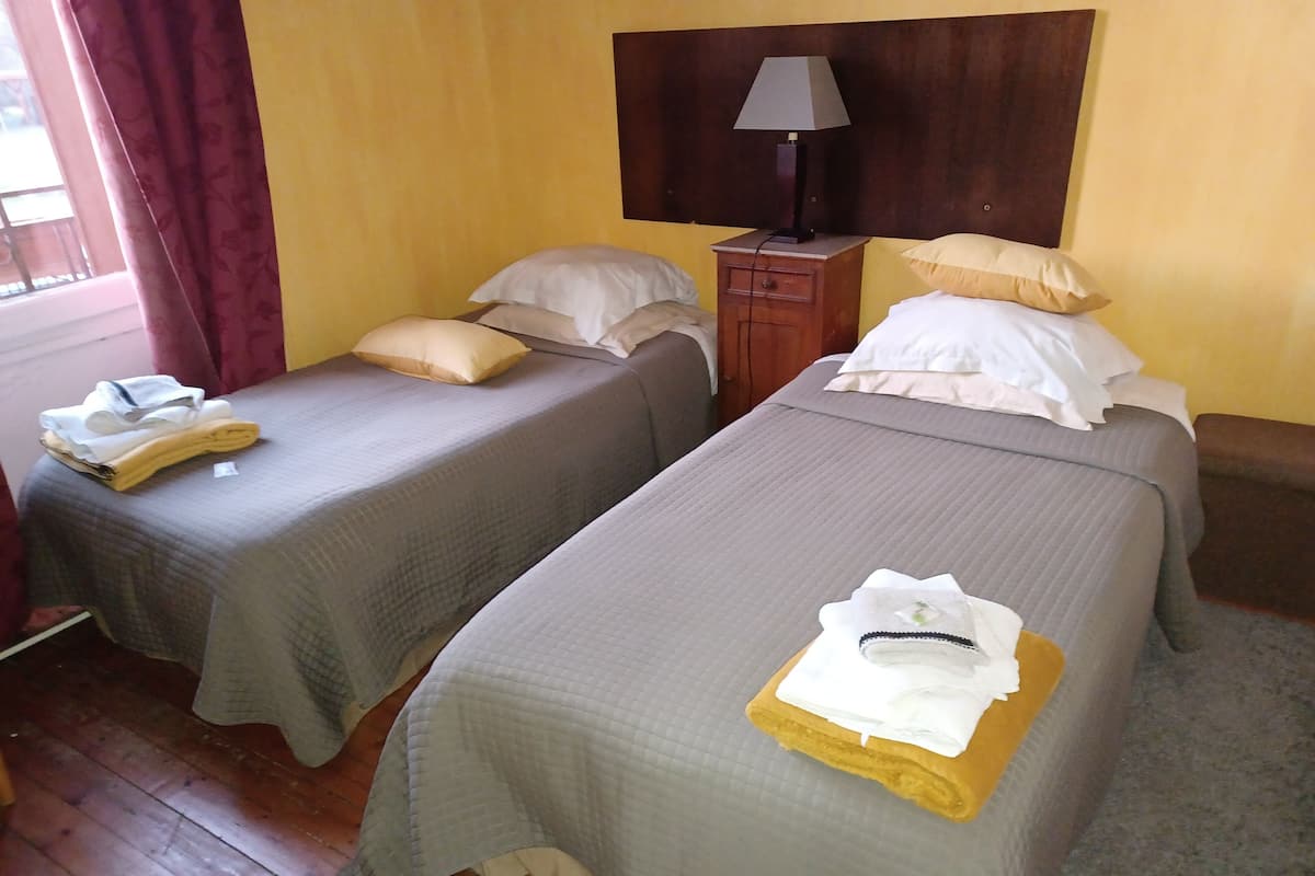 A room with two single beds, a wooden nightstand with a lamp, and a window with curtains.