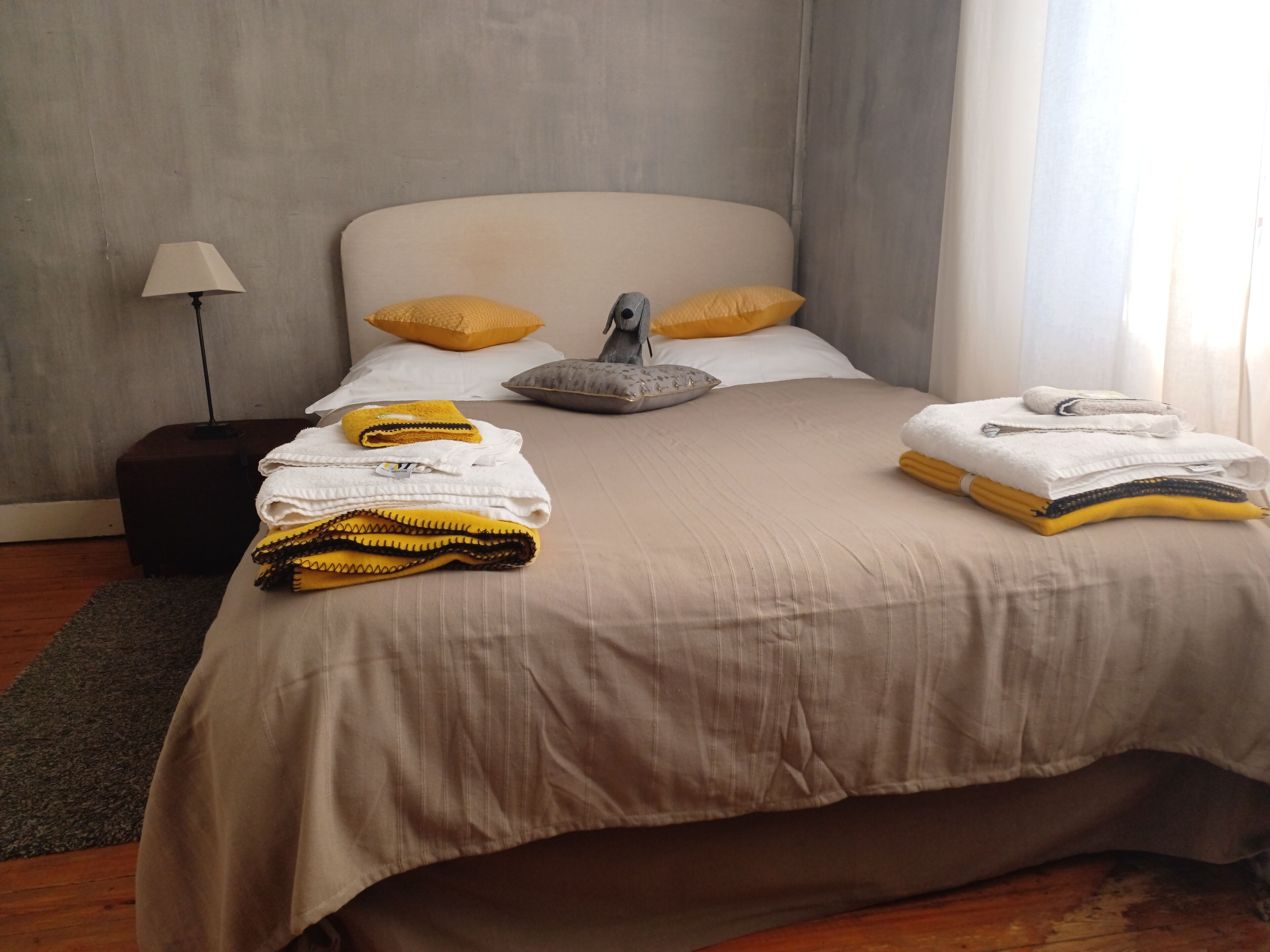 Una cama bien tendida con una colcha color beige, dos almohadas y una almohada gris sobre un piso de madera con un tapete gris.