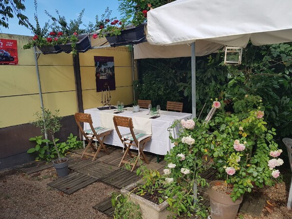 An outdoor dining area with a table set for two, surrounded by potted plants and a white canopy. - Hôtellerie Les Lilas (Bourgueil)
