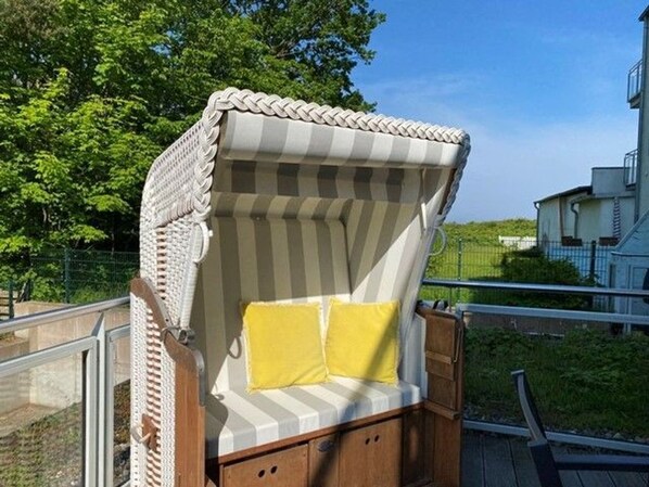 A wicker chair with a striped cushion and a yellow pillow, positioned on a wooden deck with a railing.