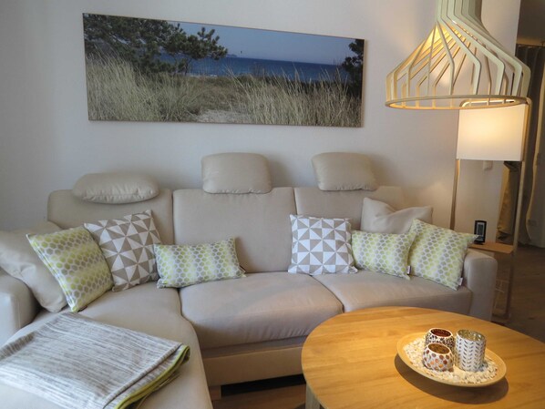 A living room with a beige sofa, patterned cushions, a painting of a beach, and a table with decorative items.