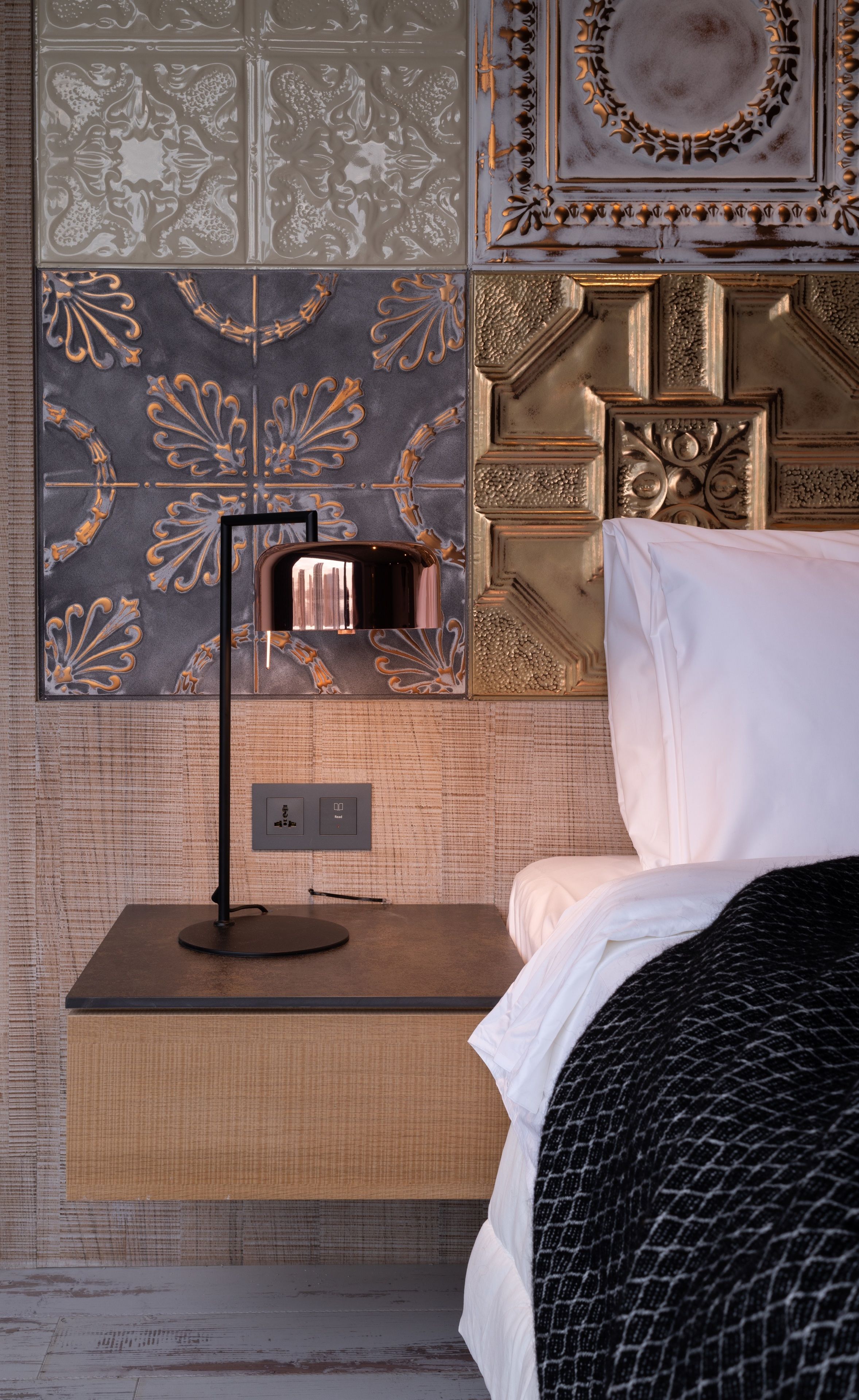 A neatly made bed with white linens, a black and white patterned blanket, and a bedside table with a lamp. The wall behind features ornate tile work and a decorative panel.