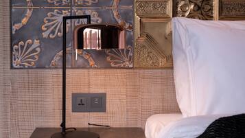 A neatly made bed with white linens, a black and white patterned blanket, and a bedside table with a lamp. The wall behind features ornate tile work and a decorative panel.