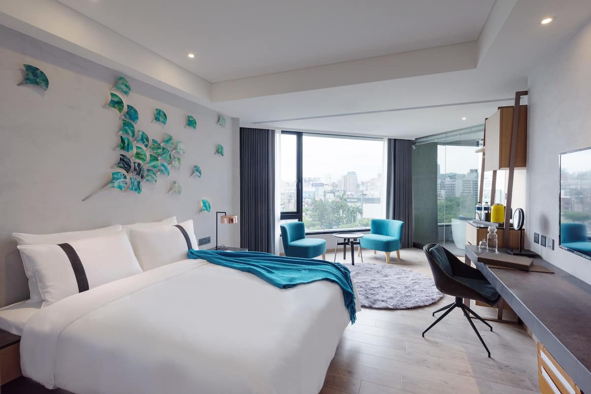 A modern hotel room with a large bed, a desk, and a view of the city.