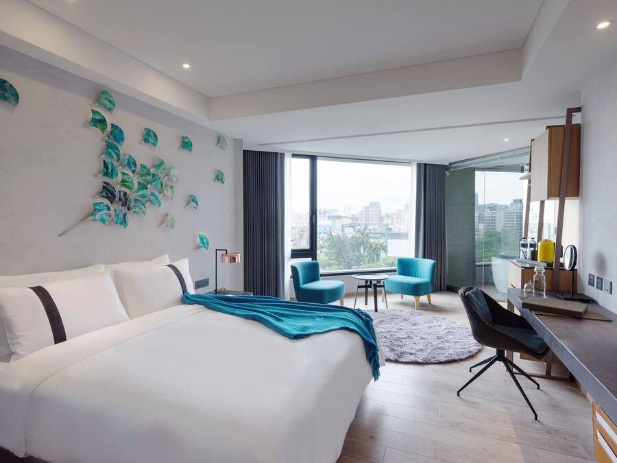 A modern hotel room with a large bed, a desk, and a view of the city.