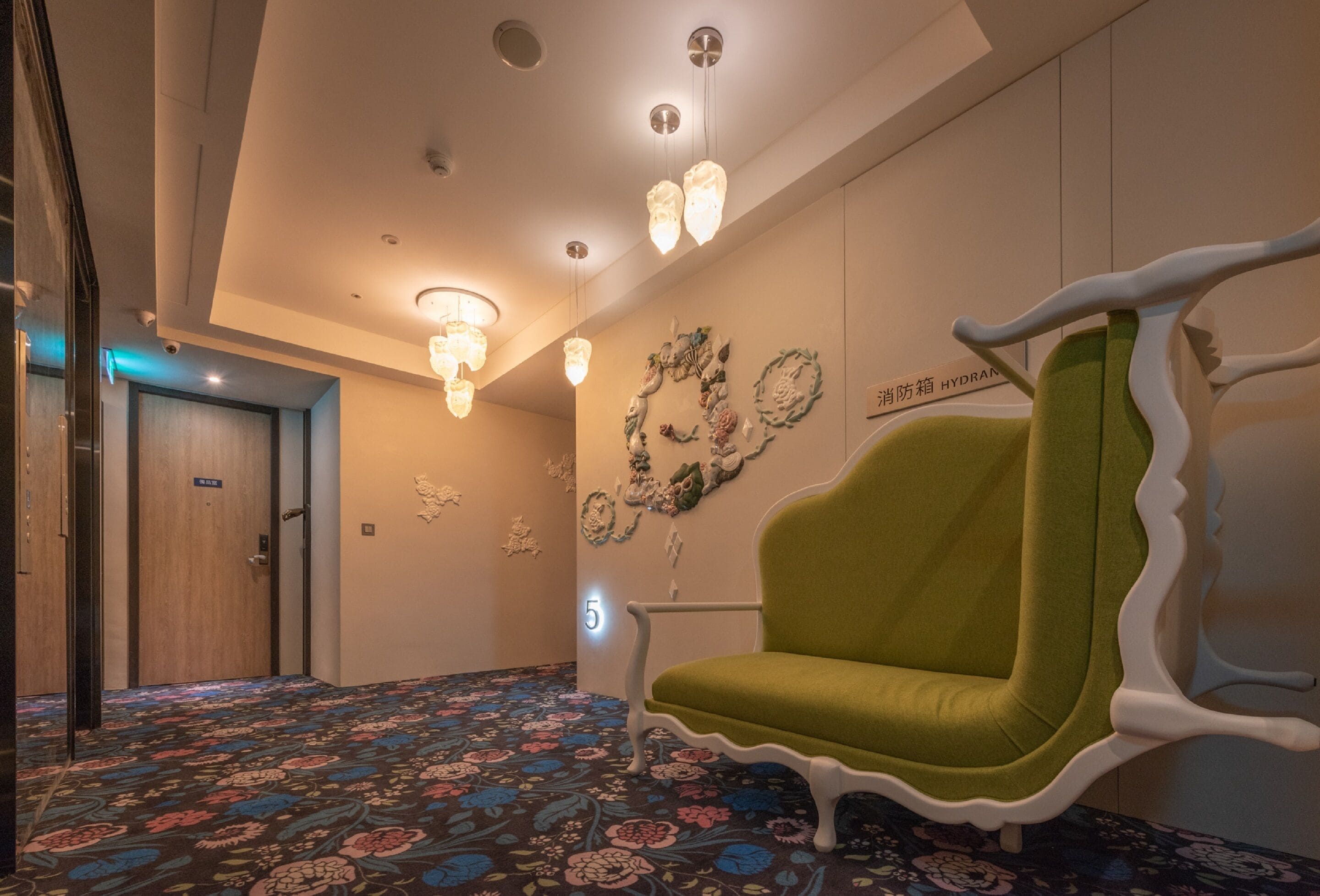 Un hall de hotel con alfombra floral, una silla verde, una pared con detalles decorativos e iluminación empotrada en el techo.