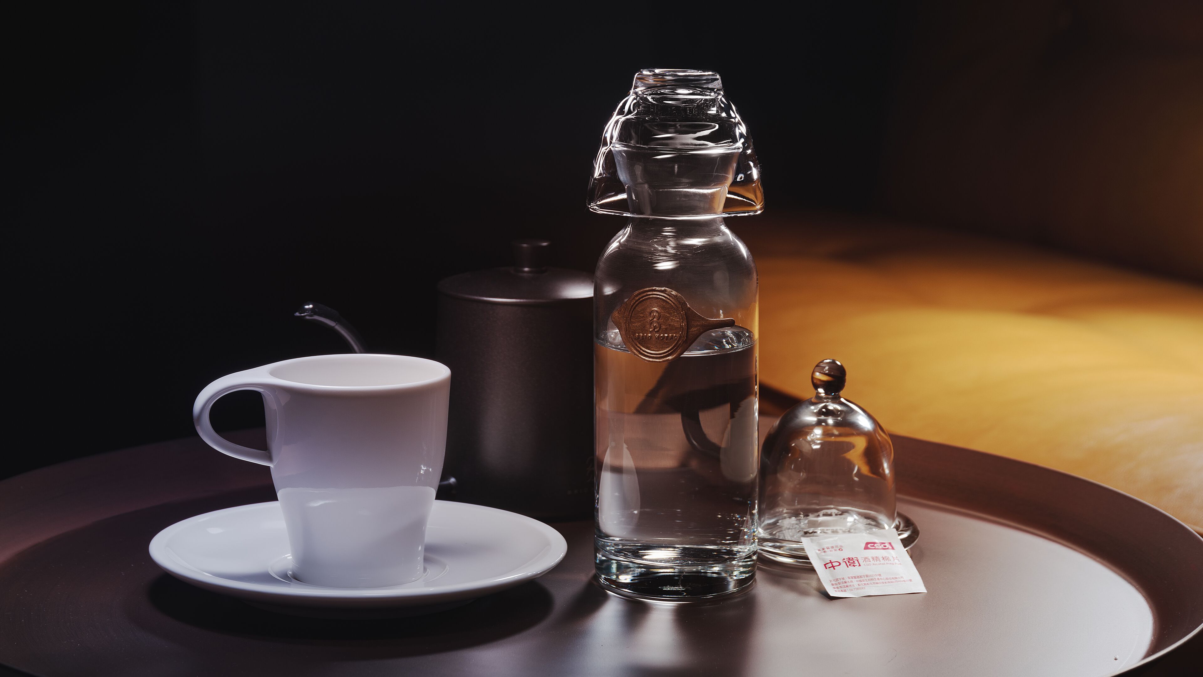 A table with a white cup and saucer, a clear bottle with a metal cap, and a small metal container with a tag.