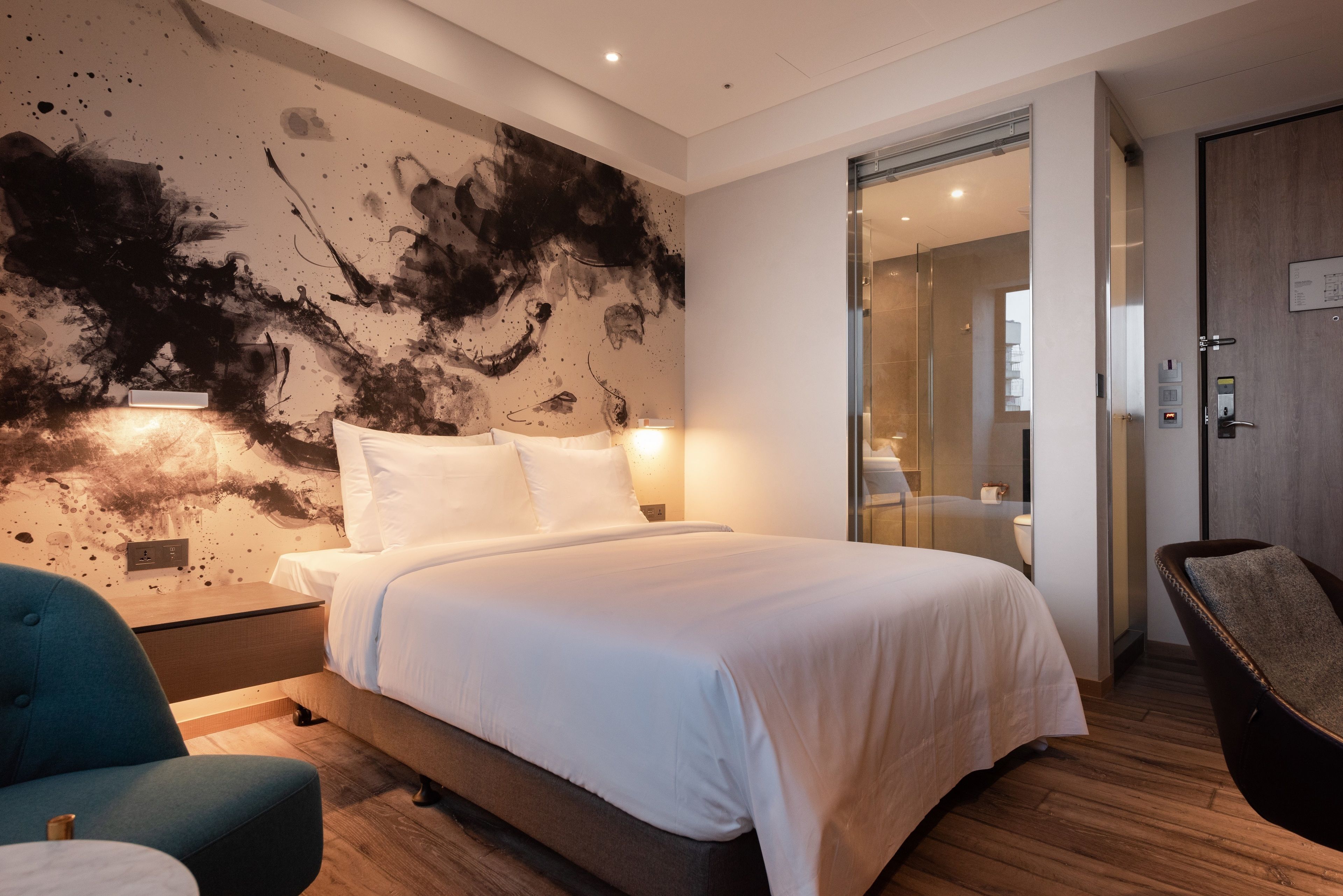 A modern hotel room with a large bed, a blue armchair, a wooden floor, and a wall with abstract art.
