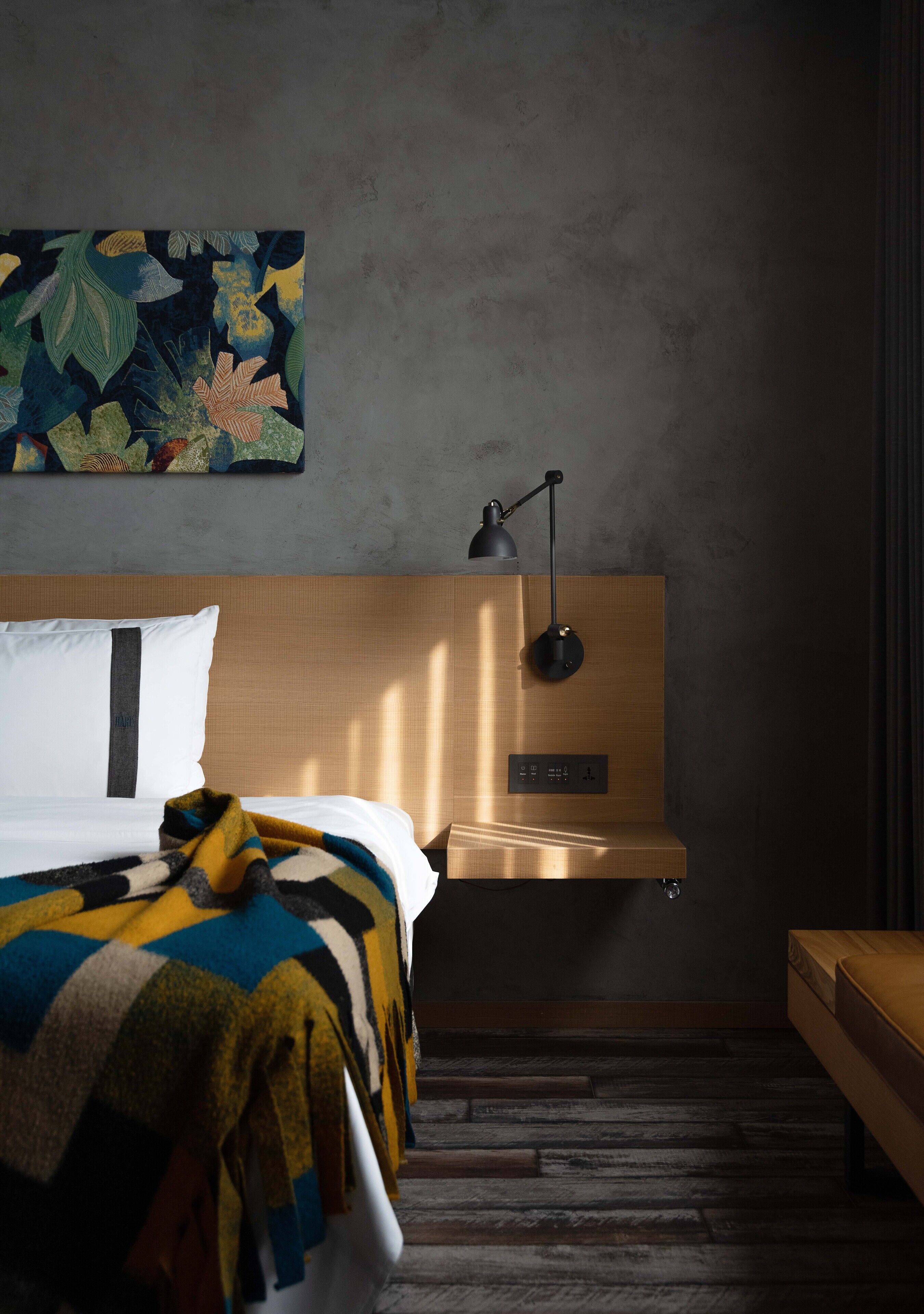 A modern hotel room with a wooden headboard, a bed with a colorful blanket, a bedside table, a desk, and a wall-mounted lamp.