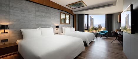Habitación de hotel con dos camas, un escritorio, una silla, un ventanal grande con vista a la ciudad y una obra de arte colgada.