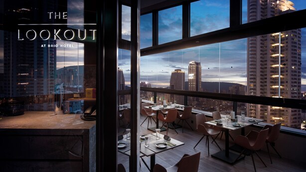 A modern hotel room with a city view, featuring a dining area with tables and chairs, and a large window offering a panoramic view of the skyline. - Brio Hotel (Kaohsiung)
