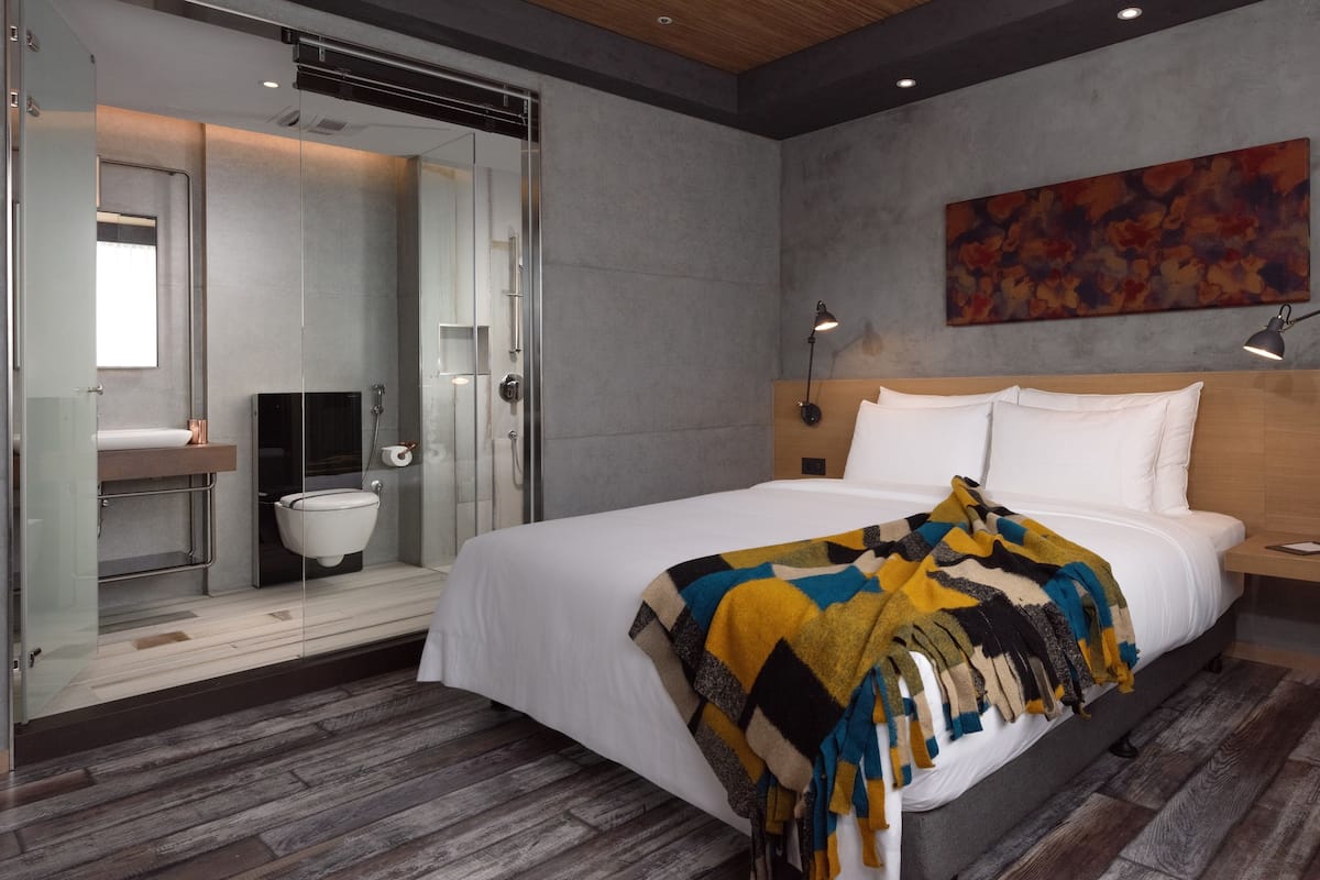 A modern hotel room with a large bed, a wooden headboard, a bedside table, a wall-mounted lamp, and a shower area with a glass door.