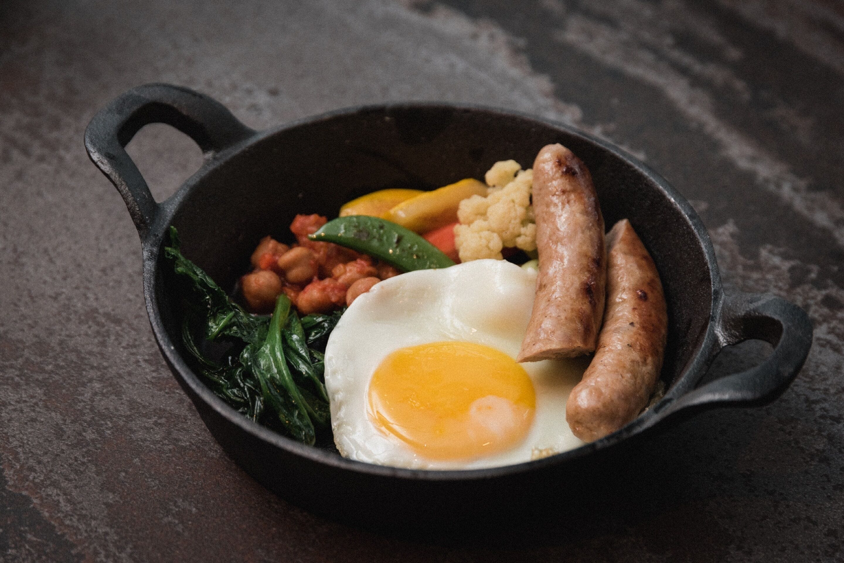 A cast iron skillet containing a fried egg, sausages, beans, and vegetables.