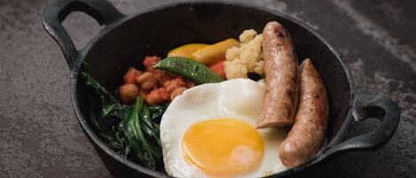 A cast iron skillet containing a fried egg, sausages, beans, and vegetables.