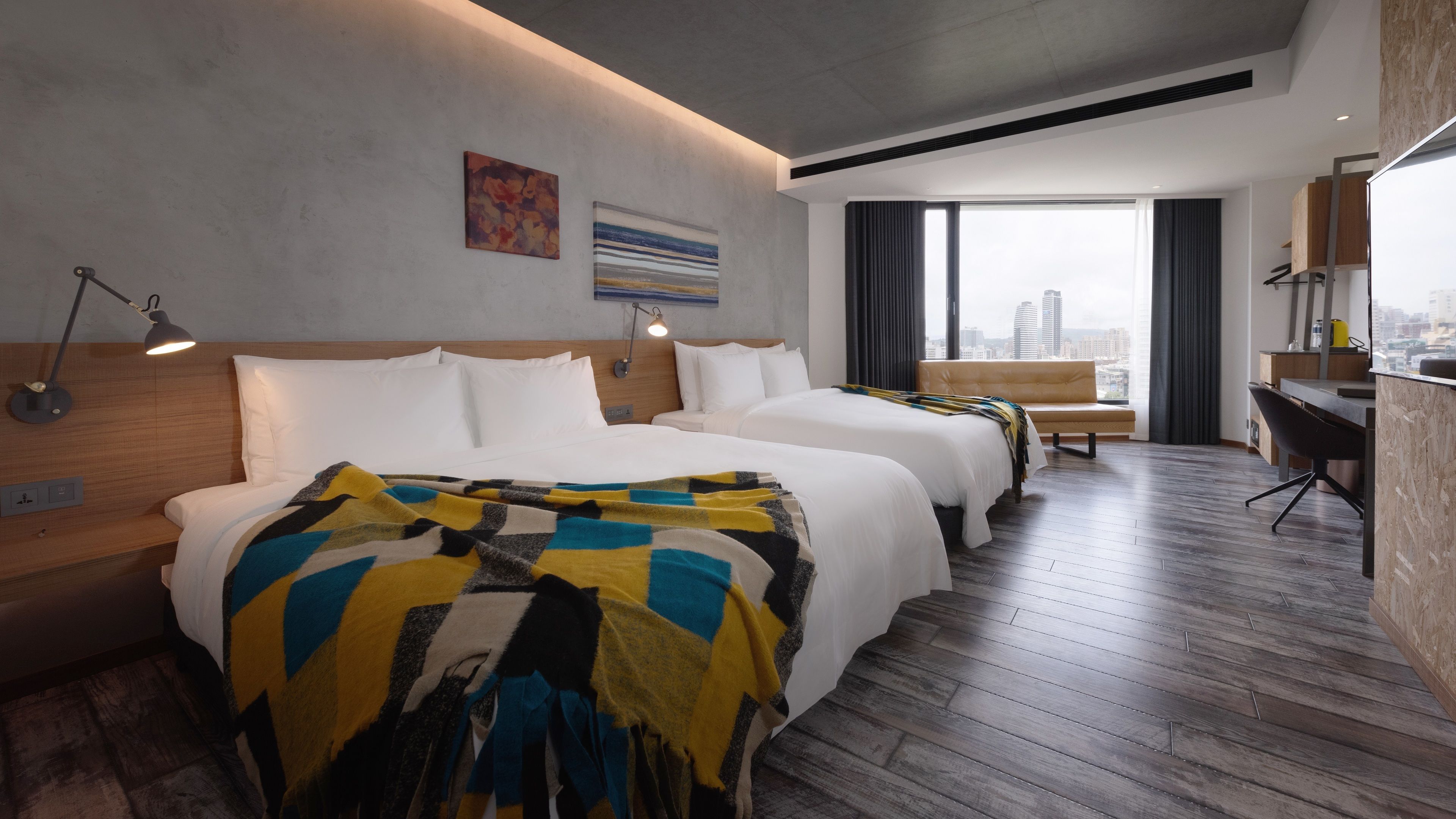 A modern hotel room with two beds, a desk, and a view of the city.