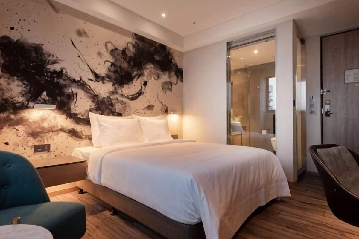 A modern hotel room with a large bed, bedside tables, a chair, and a glass partition wall.