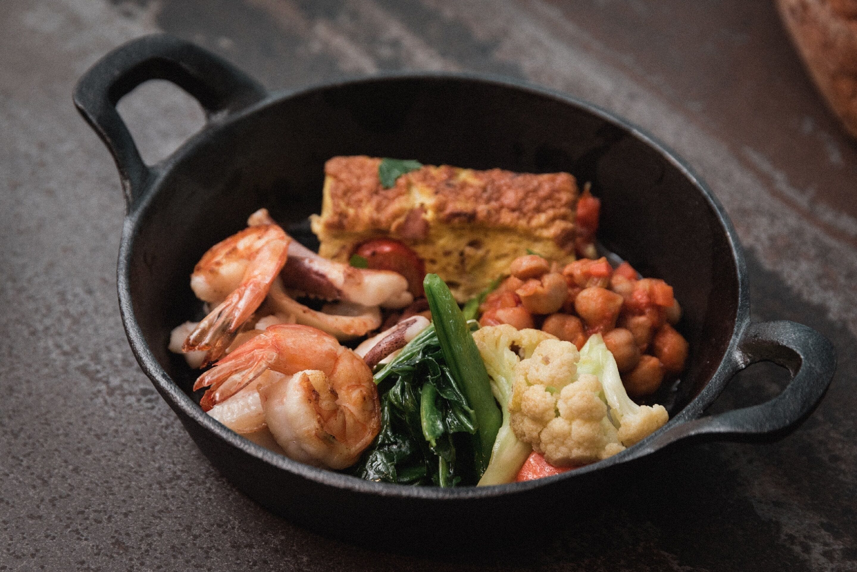 Una sartén de hierro fundido con camarones, verduras y una rebanada de pan frito.