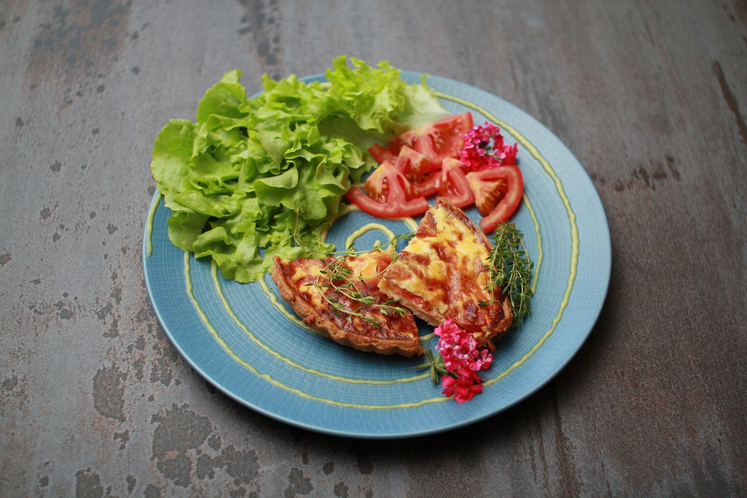 A plate with a slice of quiche, lettuce, and tomato slices on a blue plate.