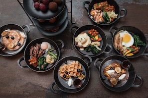 Variedad de platos servidos en sartenes de hierro fundido, como panqueques, gofres, huevos y carnes, sobre una superficie oscura.