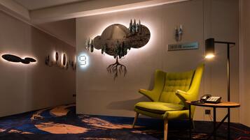 A room with a yellow armchair, a small table with a phone and a remote, and a wall-mounted artwork depicting a tree with a cloud.