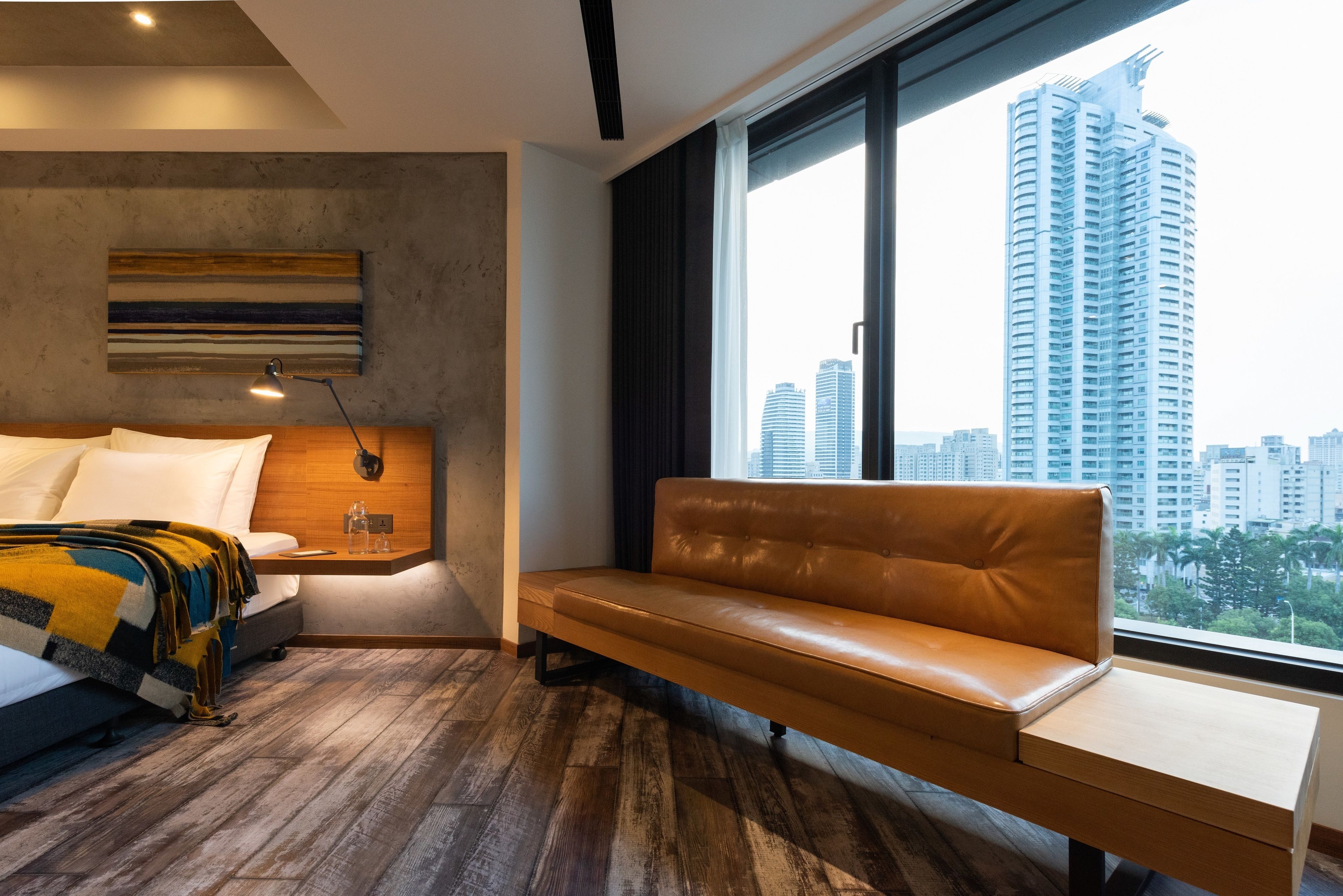 Habitación de hotel moderna con cama, banco, mesita de noche, lámpara y ventana grande con vistas a la ciudad.