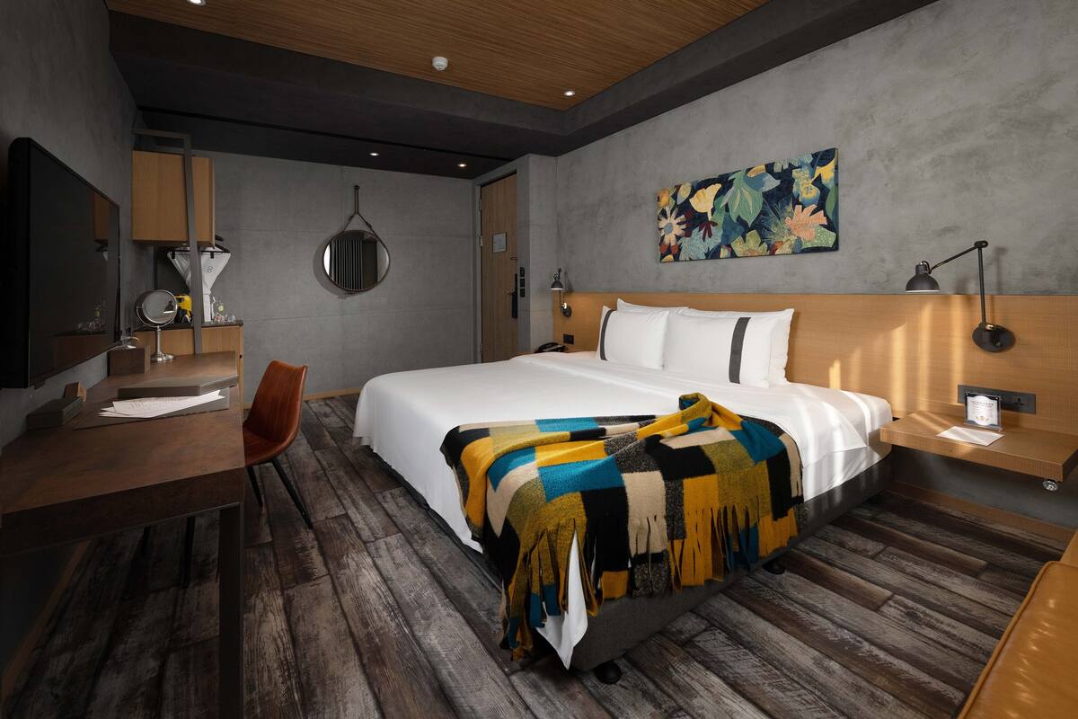 A modern hotel room with a large bed, a desk with a chair, a round mirror, a wall-mounted lamp, and a colorful artwork above the bed.
