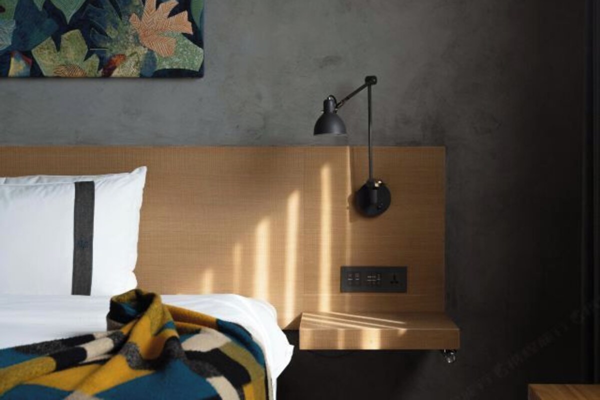 Una habitación de hotel con cabecera de madera, cama con manta a rayas, lámpara de escritorio negra y panel de control en la pared.