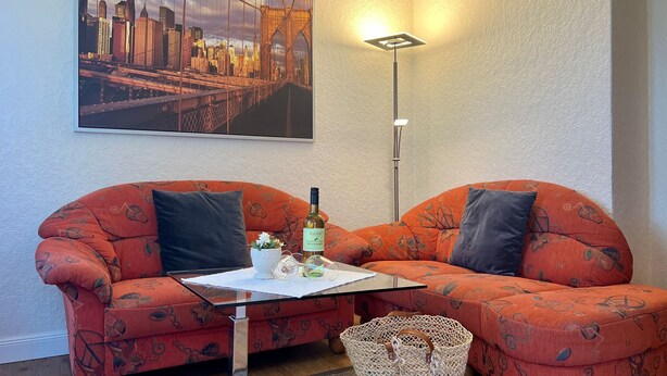 Ett vardagsrum med två orange soffor med mönster, ett soffbord i glas, en korg av rotting och en inramad tavla med en stadsbild på väggen.