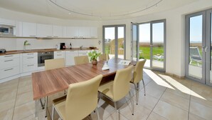 A modern kitchen with white cabinets, a wooden dining table, and chairs. There is a large window providing a view of the outdoors.