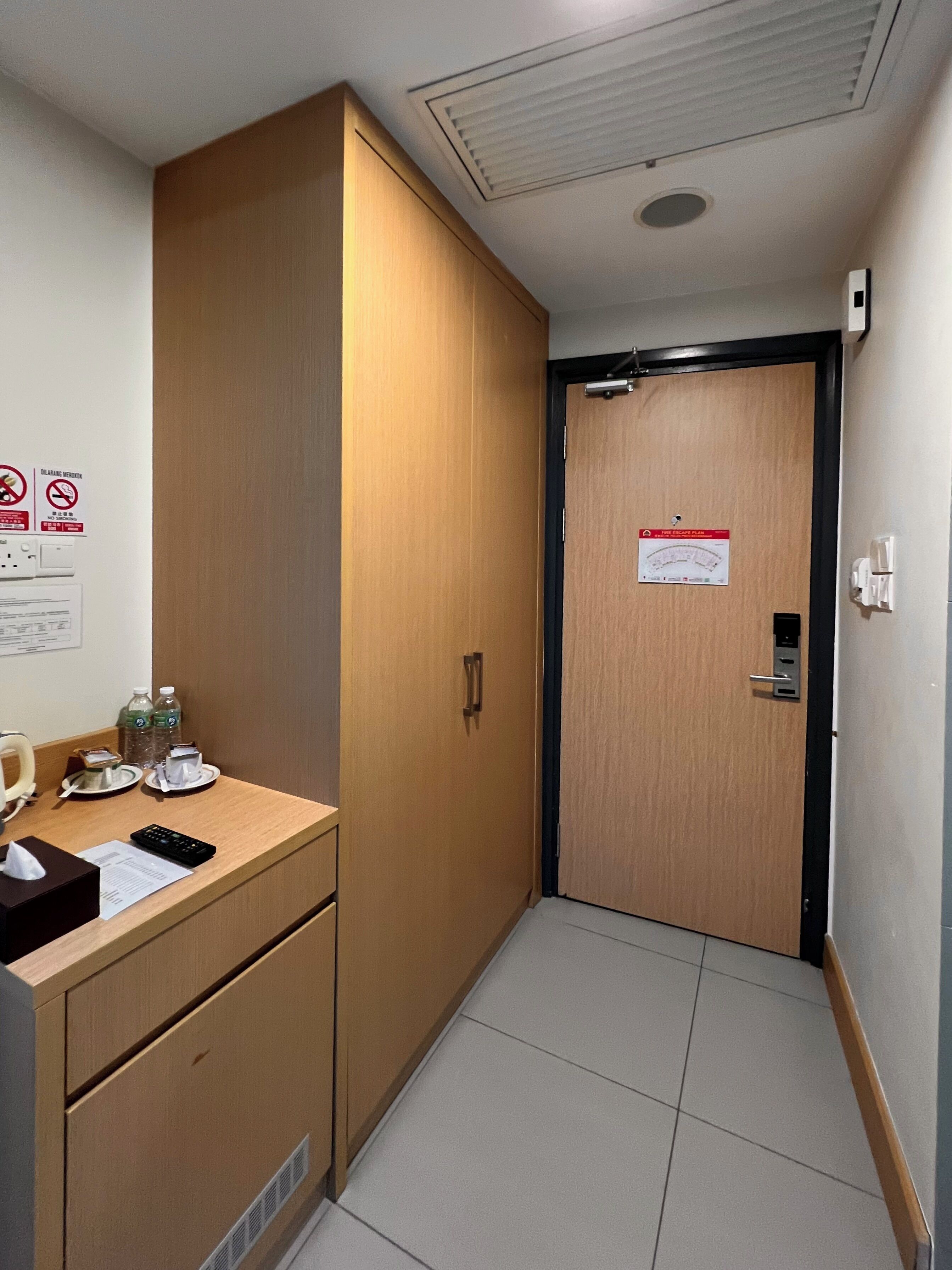 Un pasillo de hotel con un armario de madera, una pequeña mesita con artículos de aseo, un cartel de plan de seguridad contra incendios y una puerta con picaporte.