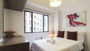 A hotel room with a large bed, a desk, a window with a view of buildings, and a painting on the wall. - Forbeswood Heights near St Lukes (Taguig)