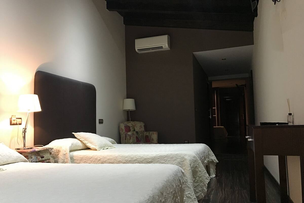 Habitación de hotel con dos camas, un escritorio, una silla, una lámpara, aire acondicionado y una puerta que da a otra habitación.