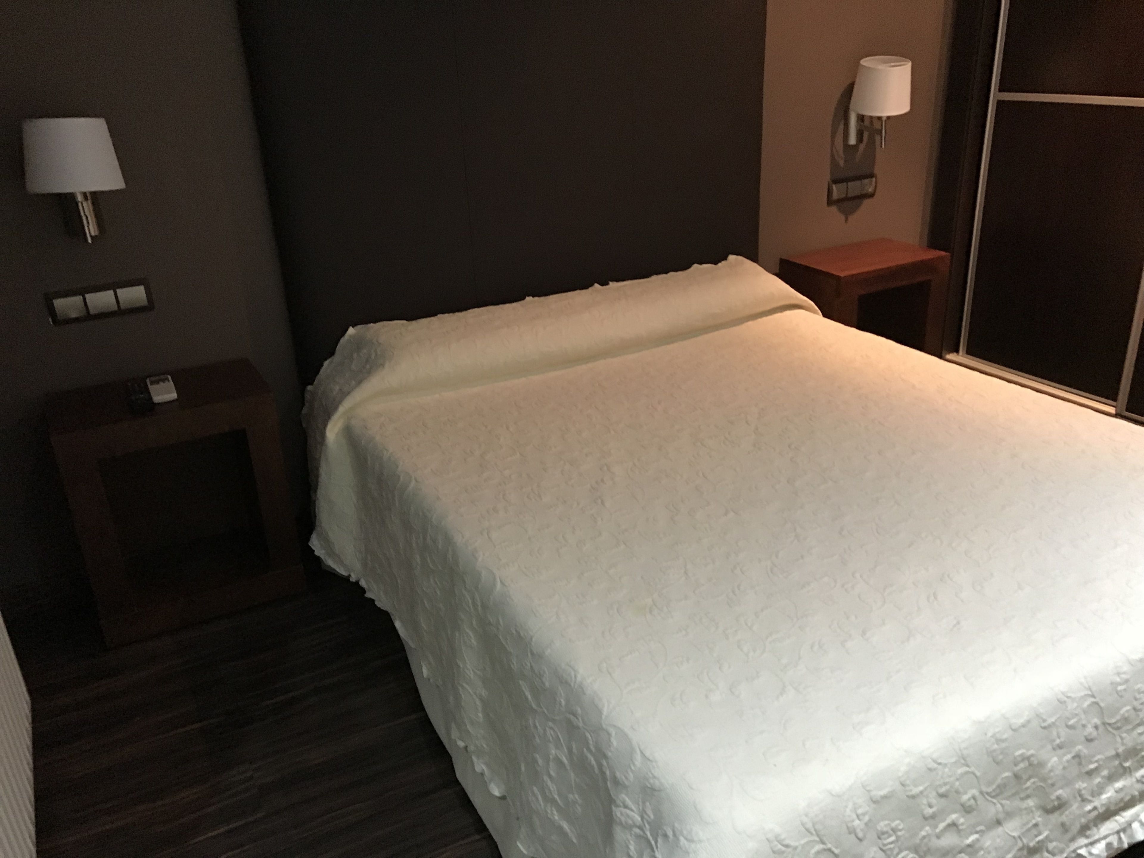 Una cama bien tendida en una habitación de hotel, con una pared oscura, una mesita de noche con lámpara y una pequeña mesita auxiliar.