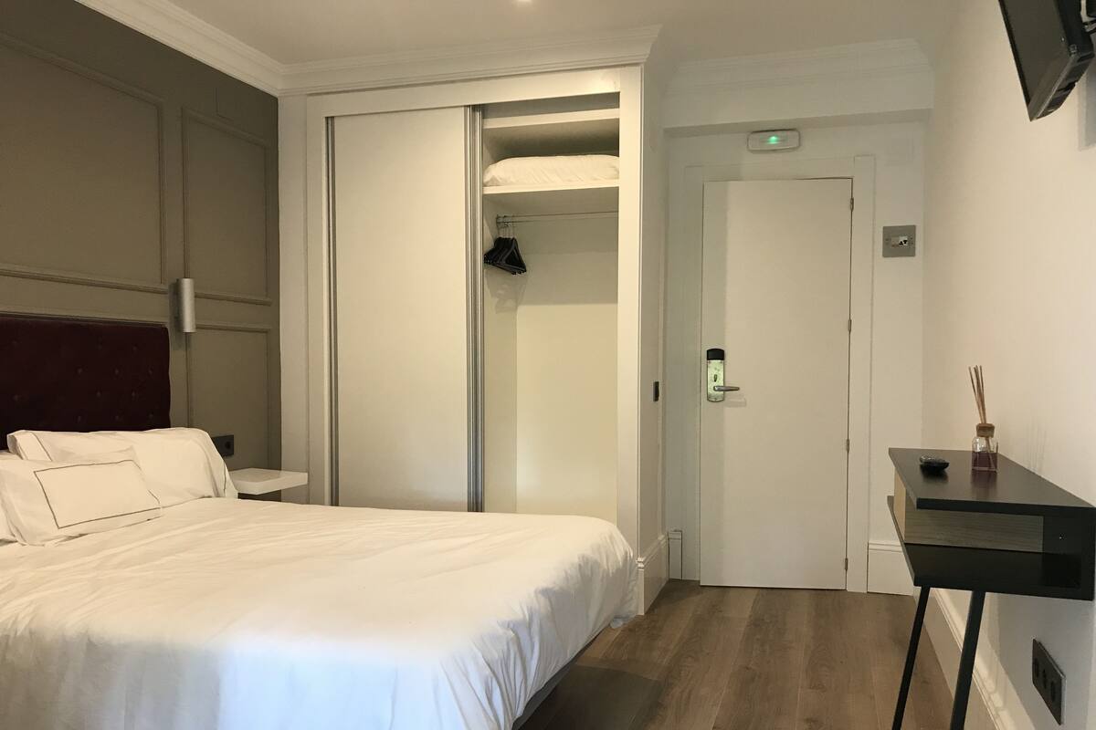 Habitación de hotel con cama, mesita de noche, escritorio con silla, armario y puerta con cerradura.