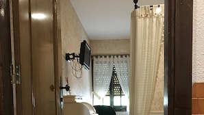 Een kamer met een houten deur, een bureau met een computer, een bed met witte gordijnen en een raam met uitzicht.