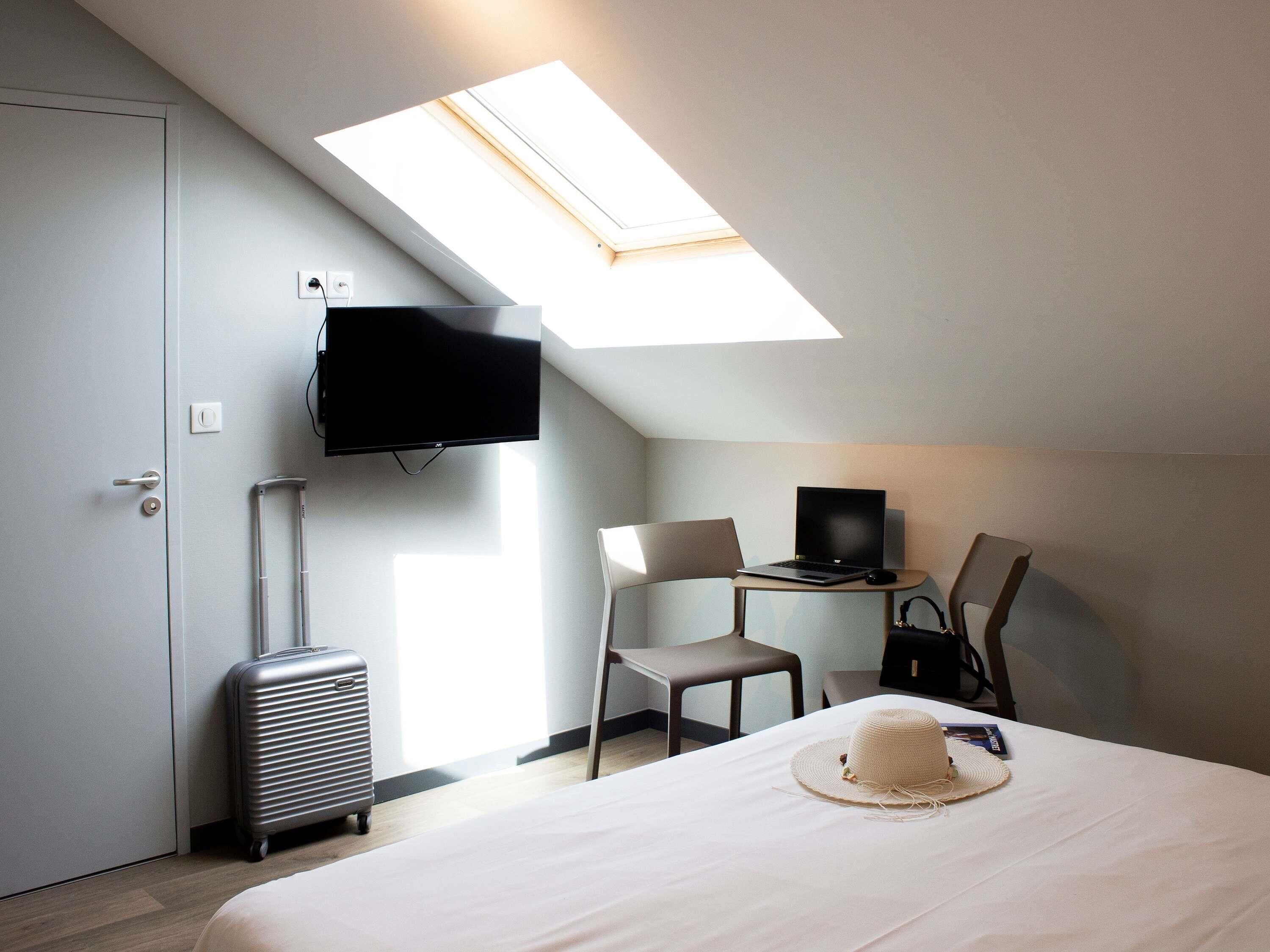 Una habitació d'hotel amb un llit, una taula de treball amb un ordinador portàtil, una televisió penjada a la paret, una maleta i un fanal.