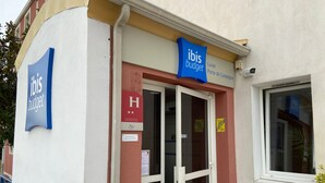 Een gebouw met een blauw ‘ibis budget’ bord, een glazen toegangsdeur en een kleine prullenbak ervoor.