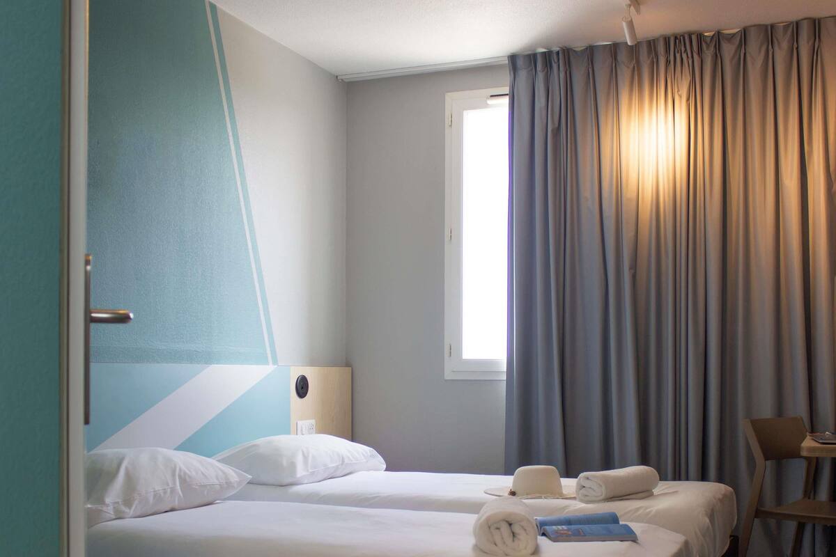 Habitació d'hotel amb dos llits, una finestra amb cortines, una taula de nit amb un barret i tovalloles, i una cadira.