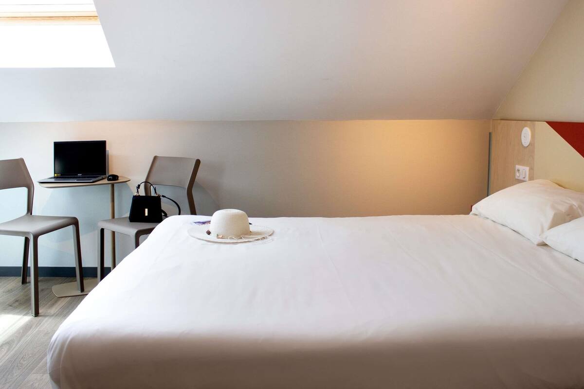 Una habitació d'hotel amb un llit, un escriptori amb un portàtil, una cadira i un barret sobre el llit.