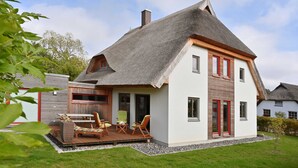 Een twee verdiepingen tellend huis met rieten dak, een houten terras met tuinmeubilair en een verzorgde tuin.