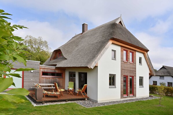 A two-story house with a thatched roof, a wooden deck with outdoor furniture, and a well-maintained garden. - Thatched house driftwood (Wiek)