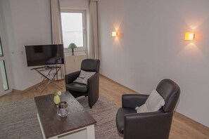 Ein Wohnzimmer mit Flachbildfernseher, zwei Ledersesseln, einem kleinen Tisch mit einer Vase und einem Fenster mit Vorhängen.