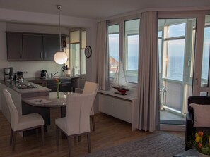 A modern kitchen with a dining area, large windows with curtains, a view of the sea, and a small sailboat decoration.
