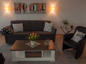 A living room with a sofa, a coffee table with a vase of flowers, a side table with a plant and a red cup, and a chair with a patterned pillow.