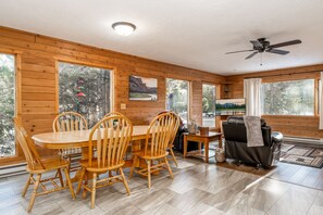 L’intérieur chaleureux d’un chalet comprend une table à manger, des chaises, un canapé, un téléviseur et de grandes fenêtres offrant une vue sur le paysage extérieur.