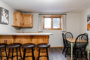 Eine Küche mit Holzausstattung, einer Bar mit Hockern, einem Esstisch mit Stühlen und einem Fenster mit Vorhängen.