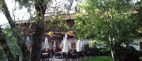 An outdoor seating area with tables and chairs, surrounded by trees and a building with a sign reading "EINGANG".