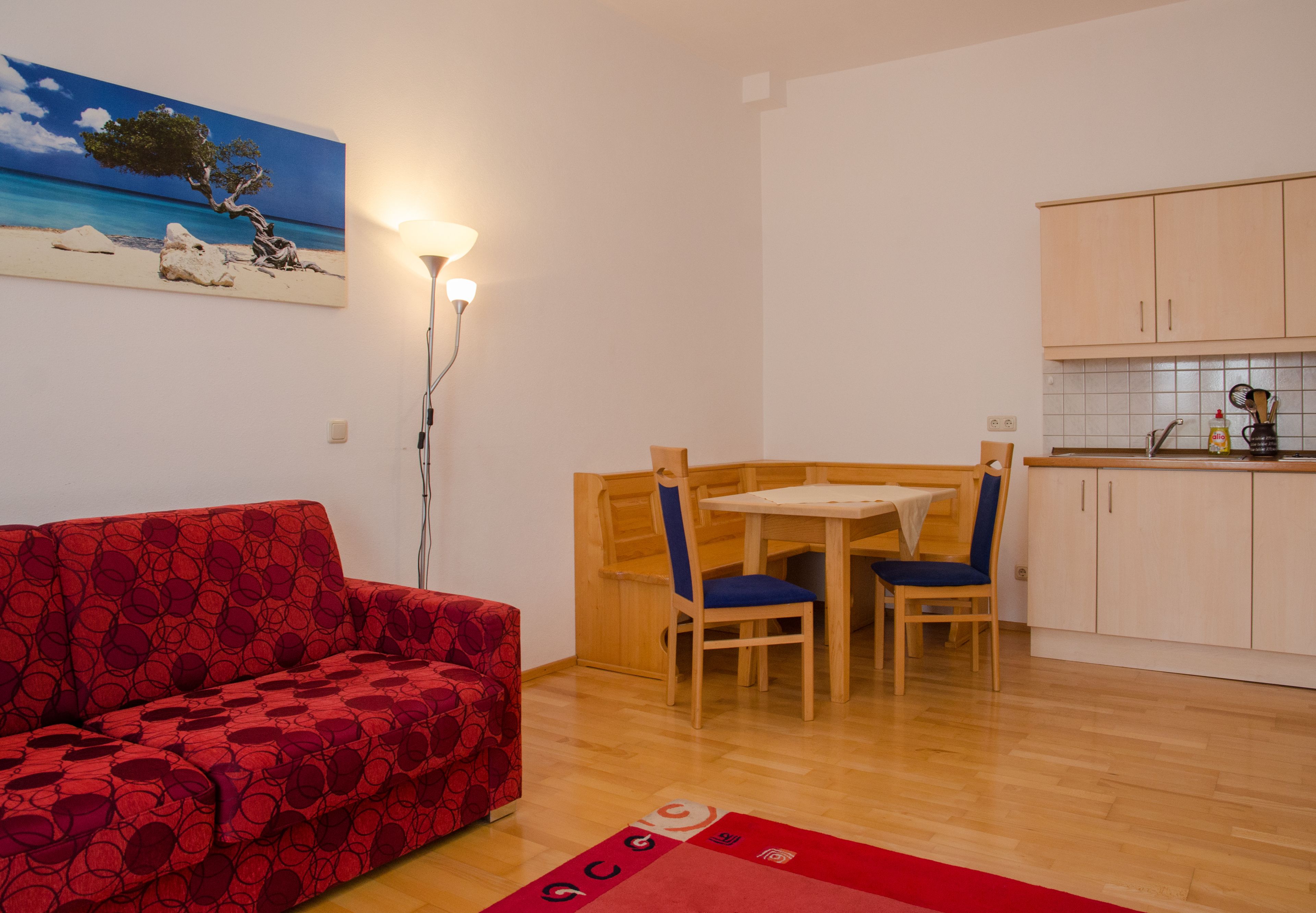 Un espacio habitable compacto con un sofá rojo estampado, una mesa de comedor de madera con dos sillas, una zona de cocina con armarios y un cuadro de un árbol en la pared.
