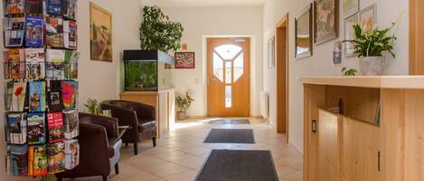A hotel lobby with a reception desk, a wall display of brochures, a potted plant, a fish tank, and a door with a geometric design.