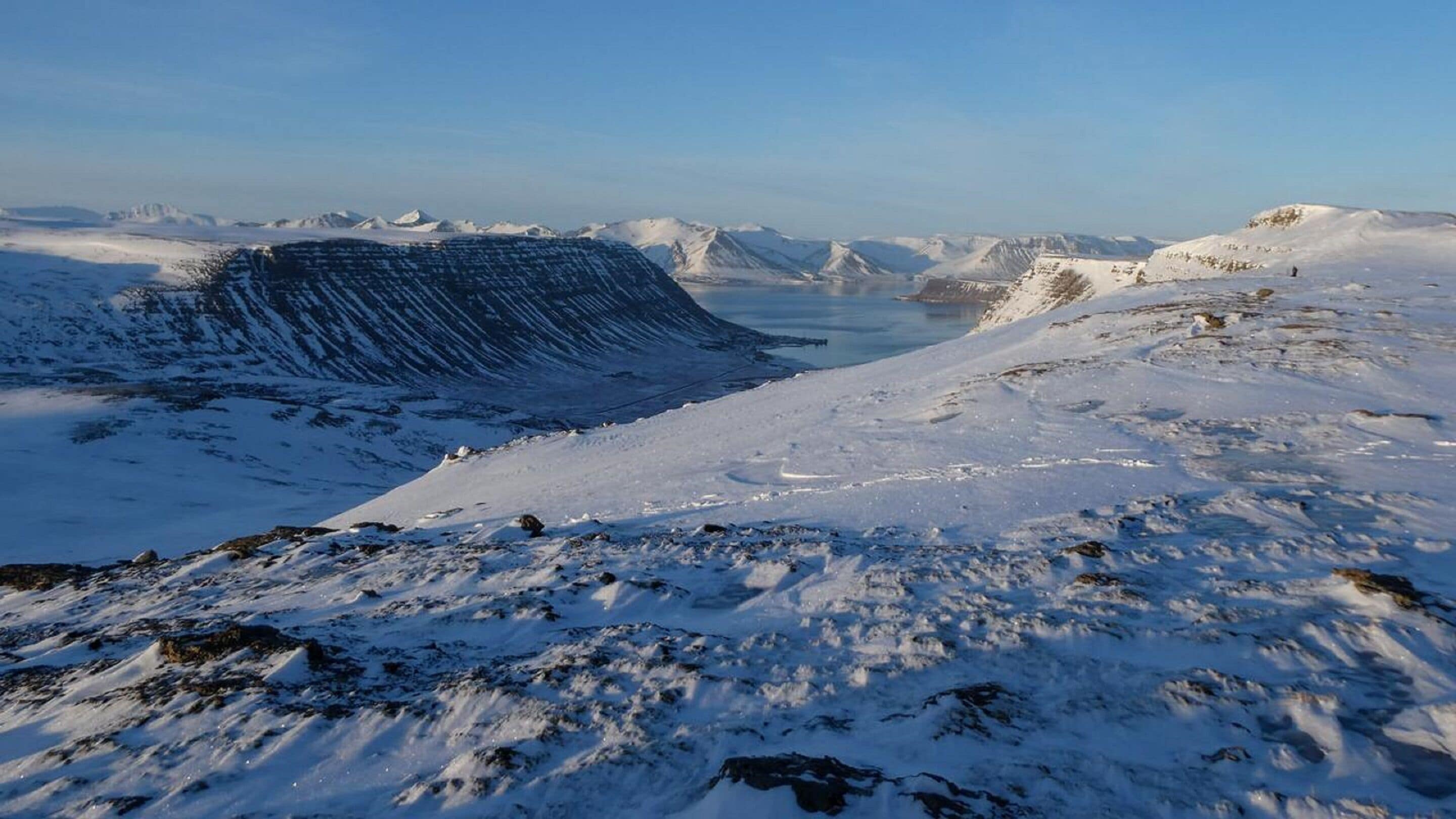 Un paisaje montañoso nevado con una cresta cubierta de nieve destacada y un lago al fondo.